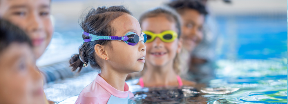 Een kind met zwembril op en haar hoofd net boven het water uit kijkt met volledige focus vooruit, om haar heen zie je verschillende kinderen lachend kijken.